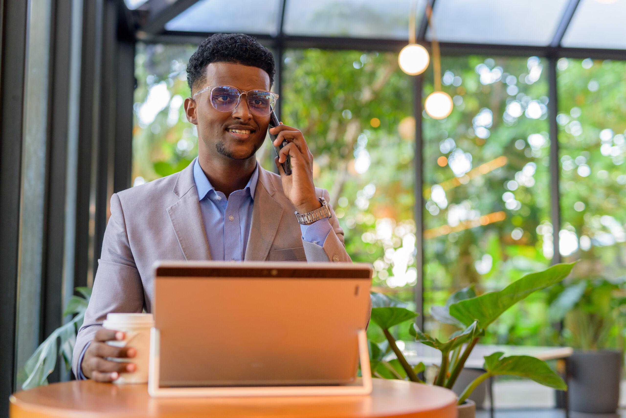 happy african businessman coffee shop using laptop computer while talking phone 1 scaled